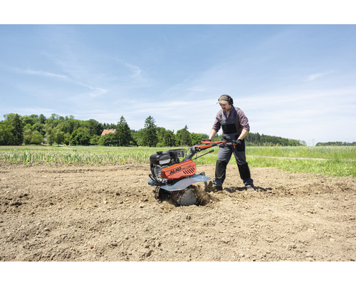 Mann arbeitet mit einer Motorhacke auf einem Feld. Im Hintergrund befinden sich Bäume und ein Haus.