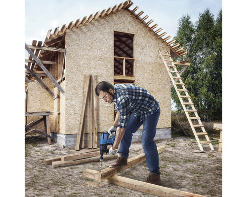 Un artisan perce une planche de bois avec une perceuse devant le gros œuvre d''une maison.