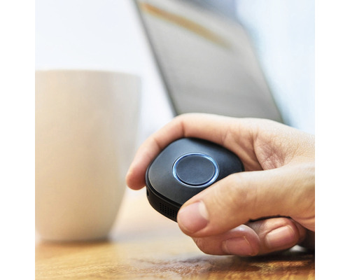 Eine Hand hält einen schwarzen Presenter mit zentralem Knopf. Im Hintergrund sind ein Laptop und eine Tasse zu sehen.