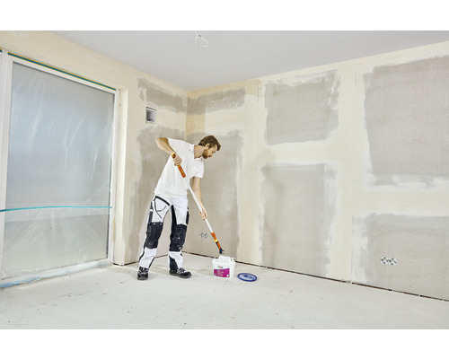 Un homme rénove une pièce avec un rouleau à peinture et un pot de peinture. Les murs sont prêts à être peints.