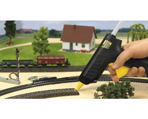 Modelleisenbahn-Szene mit Heissklebepistole in Aktion, umgeben von Miniaturhaus, Bäumen und Eisenbahnwaggons