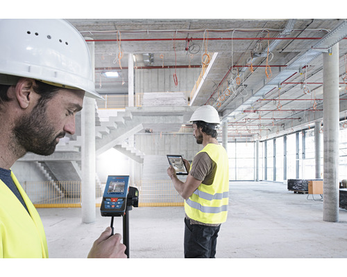 Zwei Bauarbeiter verwenden ein digitales Laserentfernungsmessgerät und ein Tablet auf einer Baustelle.