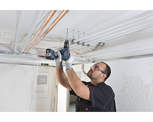 Un artisan perce des trous dans un plafond à l'aide d'une perceuse-visseuse sans fil pour installer des tuyaux et des câbles.