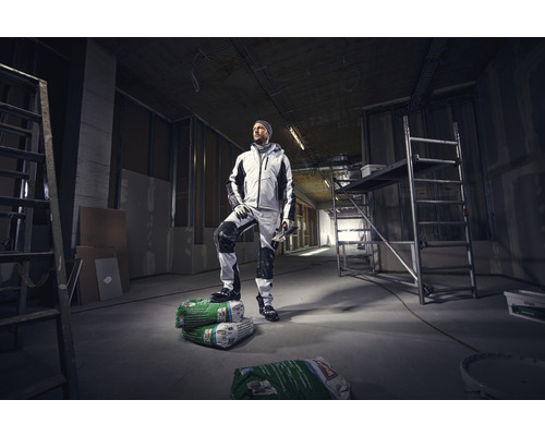 Ein Handwerker in Arbeitskleidung steht in einem Rohbau auf Säcken mit Putzmörtel.