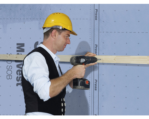 Un artisan fixe une latte en bois à un mur de maison avec une visseuse sans fil