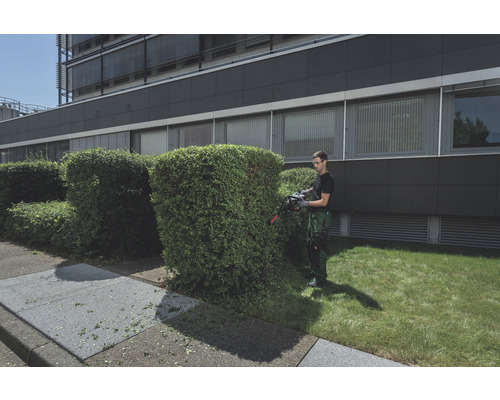 Un homme taille une haie devant un bâtiment avec un taille-haie.