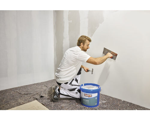 Un homme applique de lissé avec une spatule sur un mur. Au premier plan, un seau de lissé.