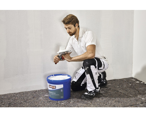 Un homme applique du mortier sur un mur à l''aide d''une spatule. Un seau de mortier est au premier plan.