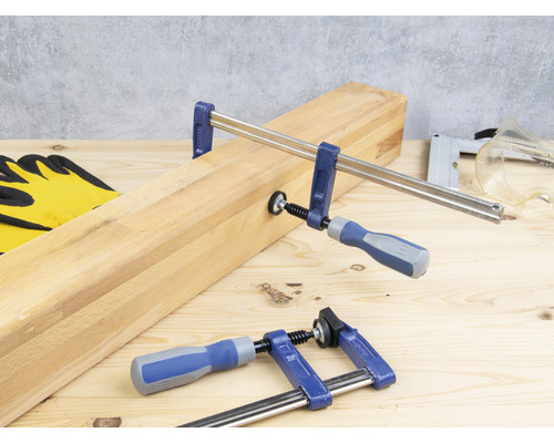 Deux serre-joints fixent une pièce de bois sur un établi, complétés par des gants de travail, une agrafeuse et des lunettes de protection.