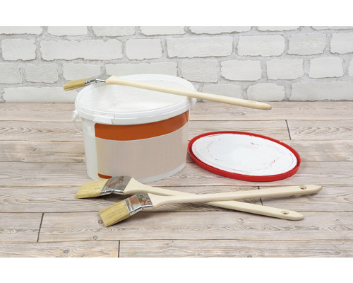 Seau de peinture, pinceaux et essoreur de peinture sur un plancher en bois devant un mur de briques blanches