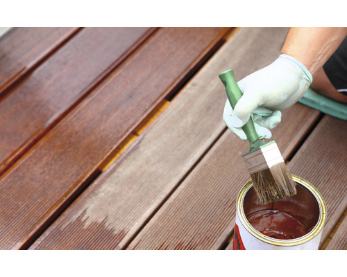 Eine Person ölt eine Holzterrasse mit einem Pinsel und einer Dose Öl.