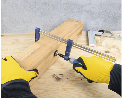 Une latte de bois est fixée avec un serre-joint à vis, actionné par des mains gantées de gants de travail.