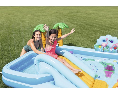 Pataugeoire gonflable avec toboggan et arroseur pour enfants dans le jardin avec deux personnes qui jouent.