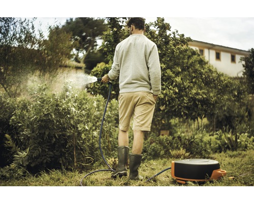 Eine Person bewässert Pflanzen im Garten mit einem Gartenschlauch und einer Schlauchbox.