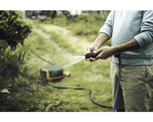 Person bewässert mit einer Gartenspritze den Rasen, im Hintergrund ein Rasenmähroboter.