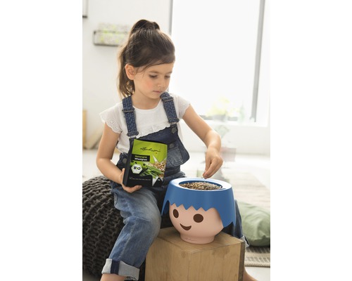 Fille remplit de graines de gazon un pot de fleurs en forme de visage, tient une enveloppe de graines, label bio