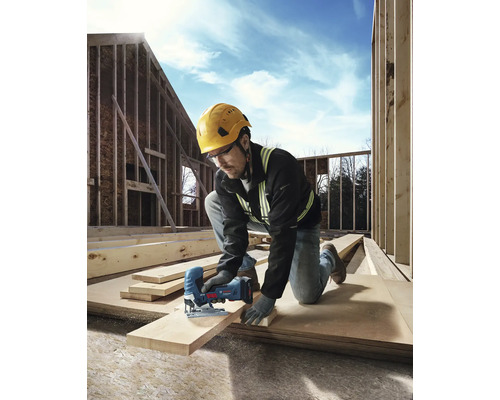 Un ouvrier du bâtiment portant un casque et des lunettes de protection est en train de scier une planche de bois avec une scie sauteuse sur un chantier.