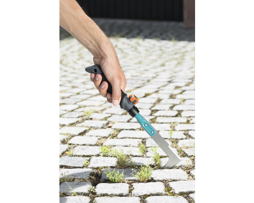 Une main tient un couteau à joints pour enlever les mauvaises herbes entre les pavés.