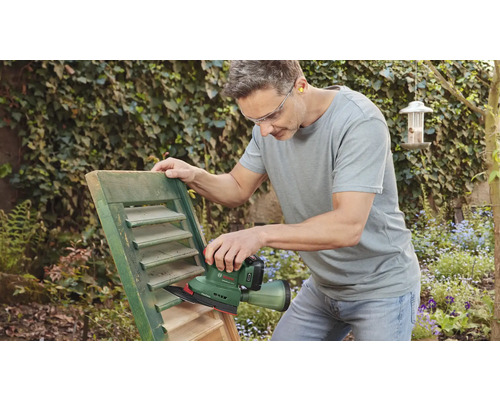 Un homme ponce un volet avec une ponceuse dans le jardin.