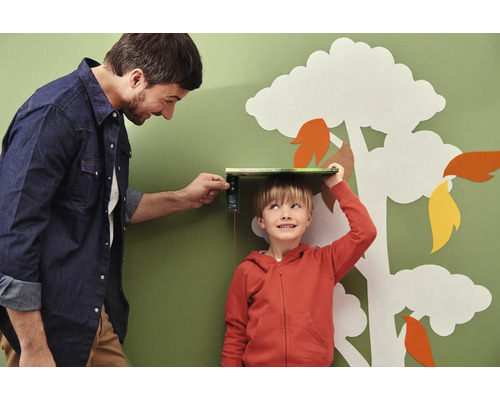 Vater misst die Grösse seines Sohnes mit einem digitalen Messgerät vor einer Wand mit Baum-Dekoration.