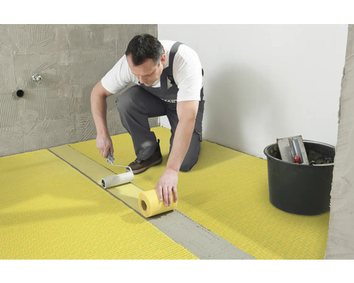 Un artisan applique une membrane d''étanchéité sur le sol à l''aide d''un rouleau à peinture. Un seau avec des outils est visible en arrière-plan.