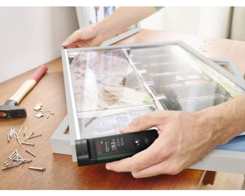 Mann misst Bilderrahmen mit Laser-Entfernungsmesser auf Werkbank mit Hammer und Nägeln