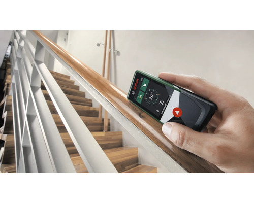 Une main tient un télémètre Bosch contre une rampe d''escalier en bois.
