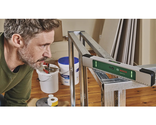Ein Mann arbeitet mit einer digitalen Wasserwaage von Bosch an einem Metallrahmen. Werkzeuge und Gipsplatten sind im Hintergrund zu sehen.
