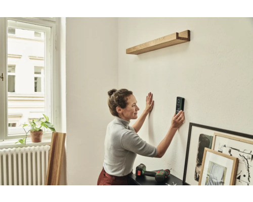 Frau benutzt ein Laser-Distanzmessgerät, um ein Holzregal an der Wand zu befestigen.