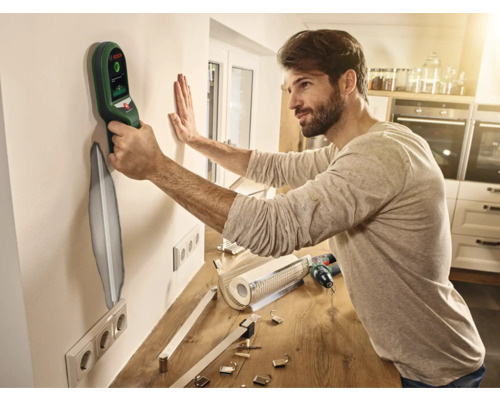 Ein Mann verwendet einen Ortungsgerät an einer Wand, um Installationen zu finden. Auf der Arbeitsplatte liegen Werkzeuge und Beschläge bereit.