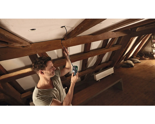 Un homme utilise un instrument de mesure pour rechercher des câbles dans un plafond, dans un grenier.