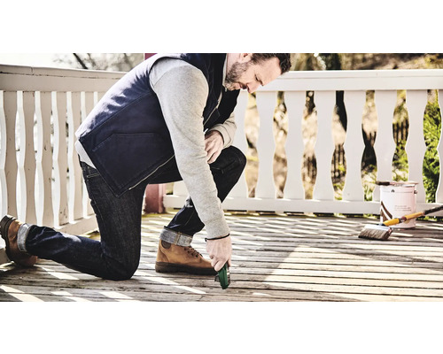 Mann kniet auf einer Holzterrasse und führt Reparaturarbeiten mit einem Werkzeug durch. Pinsel und Farbe sind im Hintergrund zu sehen.