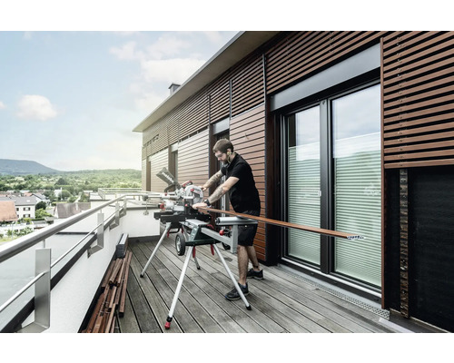 Un homme coupe une latte de bois avec une scie à onglet sur un balcon.