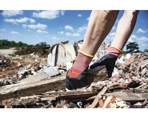 Eine Person trägt Arbeitshandschuhe und hält ein Holzstück vor einem Hintergrund aus Bauschutt.