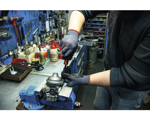 Eine Person arbeitet mit einem Schraubendreher und Schutzhandschuhen an einem Werkstück, das in einem Schraubstock befestigt ist.