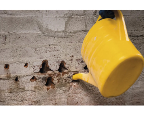 Eine gelbe Gießkanne befeuchtet eine Backsteinmauer mit Bohrlöchern zur Mauertrockenlegung.