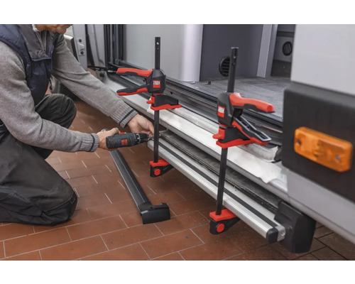 Handwerker befestigt mit einer Schraubzwinge und einem Akkubohrer ein Bauteil an einem Fahrzeug.