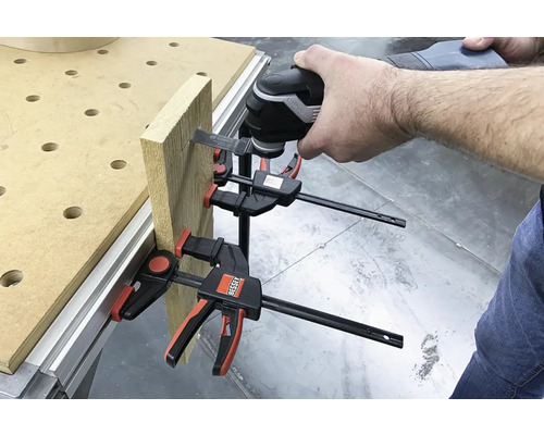 Eine Person verwendet ein Multifunktionswerkzeug an einem Holzstück, das mit Schraubzwingen an einer Werkbank befestigt ist.