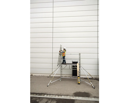 Une personne avec un casque de protection et un gilet de sécurité se tient sur un échafaudage roulant et tient une échelle contre un mur.