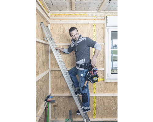 Handwerker auf einer Leiter in einem Rohbau mit Holzverkleidung und Werkzeugtasche