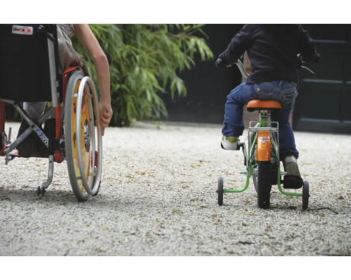 Ein Kind im Rollstuhl und ein Kind auf einem Dreirad fahren auf einer Schotterfläche.