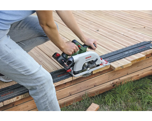 Eine Person sägt ein Holzbrett mit einer Handkreissäge auf einer Holzterrasse.