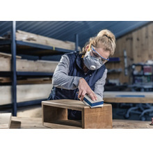 Frau schleift Holzbox mit Schleifklotz. Sie trägt Schutzbrille und Atemschutzmaske.