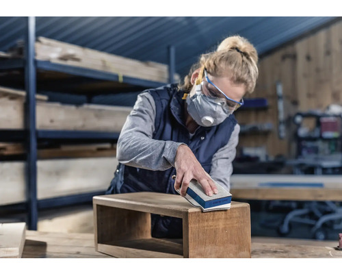 Une femme ponce une boîte en bois avec un bloc de ponçage. Elle porte des lunettes de protection et un masque respiratoire.