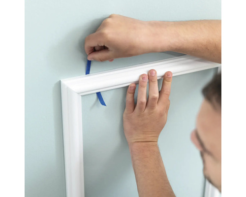Person befestigt weisse Zierleiste mit Klebeband an der Wand.
