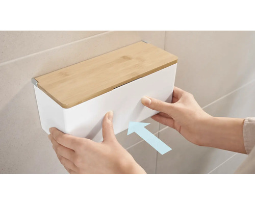 Eine Person montiert einen weissen Kosmetiktuchspender mit Bambusdeckel an der Wand.