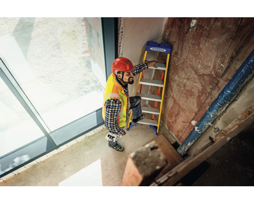Un homme avec un casque de chantier et un gilet de sécurité monte un escabeau pliant à l''intérieur.