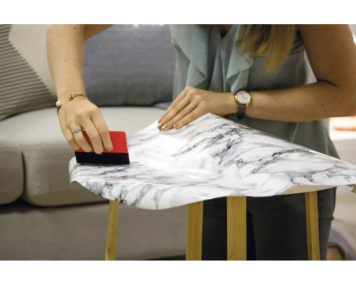 Une femme applique un film autocollant imitation marbre sur un tabouret en bois à l''aide d''une raclette