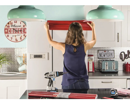 Frau befestigt Möbelfolie in Küche. Im Vordergrund Akku-Bohrschrauber und Rollen der Folie.