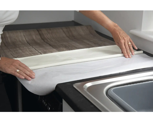 Une personne pose un film pour meubles imitation bois sur un plan de travail de cuisine.